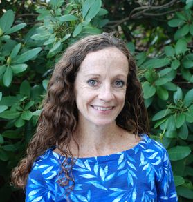Headshot of WVU professor Charlene Kelly. She is pictured against a background of green bushes wearing a blue floral top. She has long, auburn-colored curly hair. 