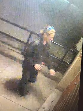 A surveillance photo shows a person in all black with blond hair approaching a building.