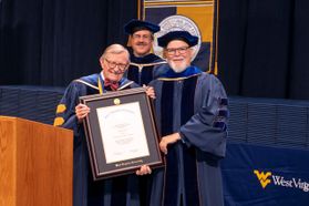 WVU President Gordon Gee hands a framed honorary degree to Larry Groce. Both are wearing navy blue academic regalia. Behind them in a matching navy blue gown and cap is Scott Wayne, Faculty Senate chair.