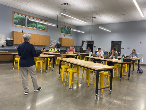 WVU Professor Frankie Tack, wearing a dark, long-sleeve shirt, khaki pants, and short gray hair, prepares WVU Extension faculty, seated three to a table on bright yellow stools, to give workshops. 