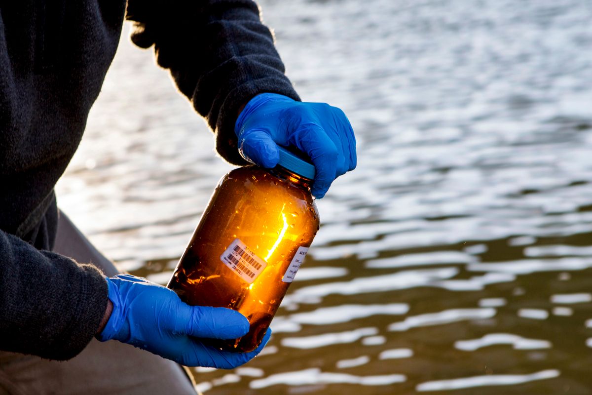 hands in blue gloves hold a brown jar with bar code in front of a body of water