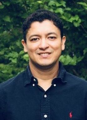 Headshot of WVU professor Carlos Quesada. He is pictured outside standing in front of green bushes. he is wearing a navy blue button up dress shirt and has short, black hair. 