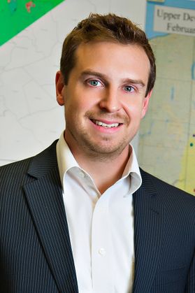 Headshot of Northwest Natural Energy executive BJ Carney. He is pictured inside standing in front of a map. He is wearing a pinstriped sport coat over a white dress shirt. He has short light brown hair and a 5 o'clock shadow. 