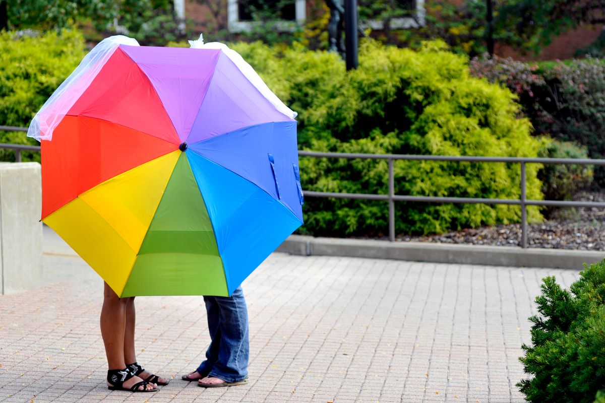 Two people stand behind a rainbow umbrella. Only their legs are visible on a sidewalk surrounded by a railing and bushes.