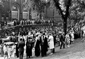 people in commencement regalia stand outside a brick building
