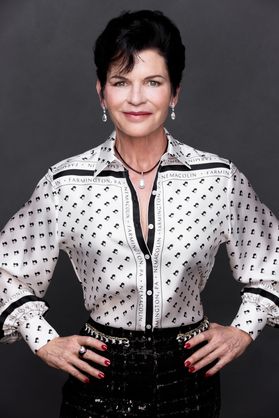 Headshot of Maggie Hardy. She is standing with her hands on her hips in front of a gray background. She is wearing a black and white patterned blouse with black bottoms and a black necklace. She has short, black hair. 