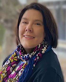 Headshot of WVU nursing professor Angel Smothers. She is pictured outside with a blurred background. She has short brown hair and is wearing a navy blue top with a pink, white, yellow and purple floral scarf around her neck. 