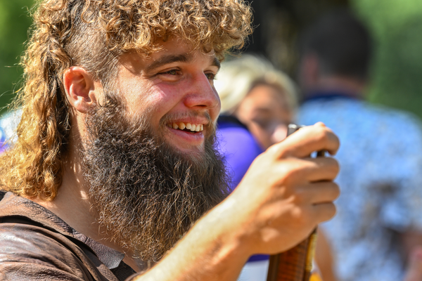 The WVU Mountaineer Mascot, Mikel Hager, is pictured here wearing his traditional buckskins and holding his rifle. He has long curly light brown hair cut in a mullet style and a long brown beard. 