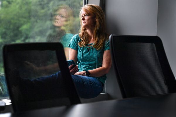 A person with long blonde hair wearing a teal shirt sits in a conference room while leaning against a window behind two black chairs.