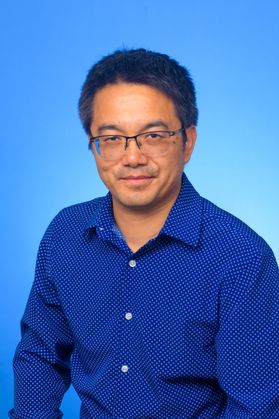 WVU researcher Michael Hu. He is pictured against a bright blue background and is wearing a royal blue dress shirt. He has short black hair and wears glasses. 