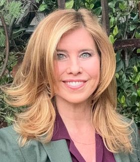 Headshot of Hollywood screenwriter Dawn DeNoon. She is standing in front of green bushes wearing a sage colored jacket over a burgundy blouse. She has long, blonde hair with layers. 