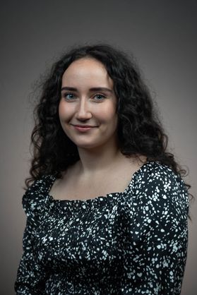 Headshot of WVU AmeriCorps VISTA Carolina Rascon. She is pictured against a dark gray background wearing a black and white patterned blouse. She has long, curly black hair. 