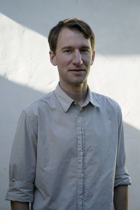 An upper body shot of a thin gentleman not smiling wearing a grey shirt.