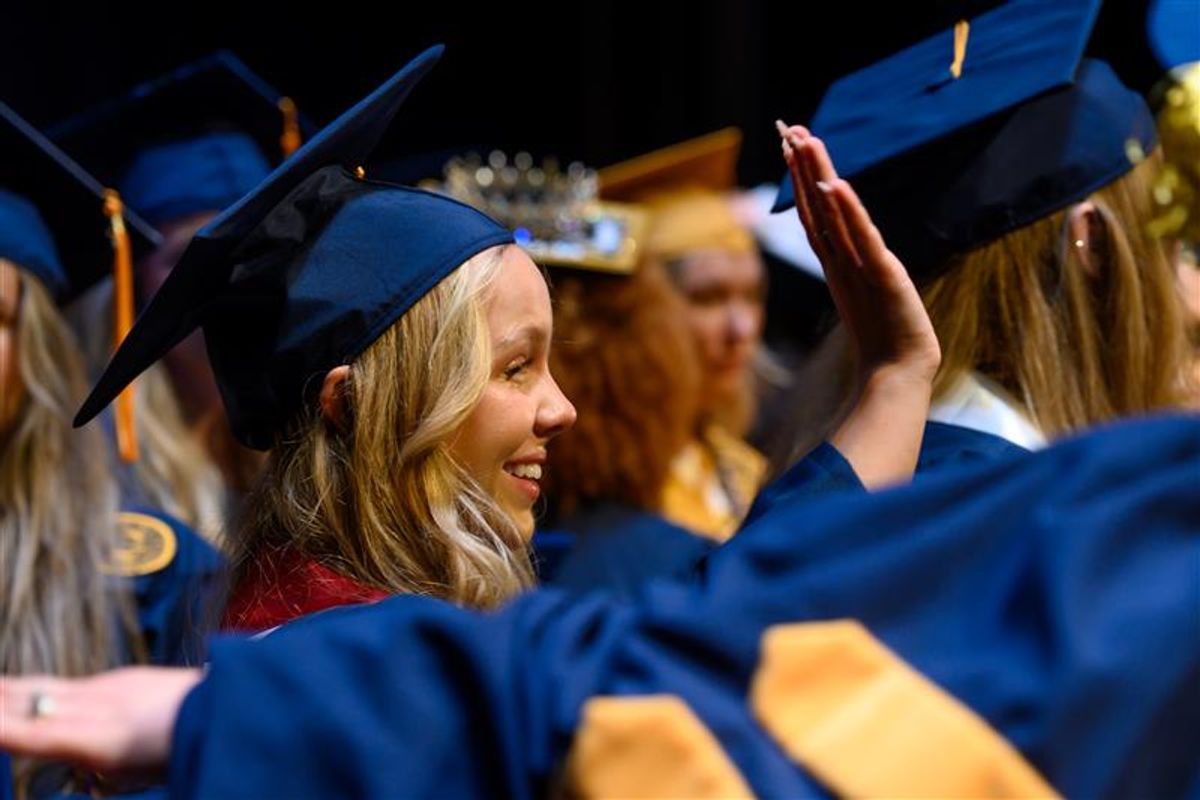 Photo of WVU graduates dressed in their navy blue and gold regalia during a Commencement Ceremony. A blonde, female graduate is in the center of the photo and is waving. 