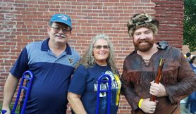 An picture of alumni Amy and Donald Hall with the Moountaineer mascot. Donald Hall is wearing a navy blue golf shirt and blue baseball cap. Amy is wearing a navy blue WVU shirt and has long gray hair. The Mountaineer is wearing his buckskins. 