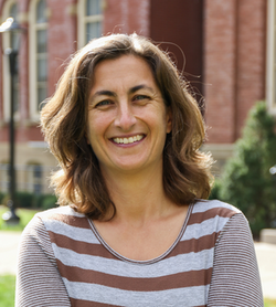 Headshot of Amy Hessl, director of the WVU Office of Undergraduate Research. She is pictured outside with a red brick building and bushes in the background behind her. She is wearing a maroon and gray striped shirt and has shoulder length light brown hair