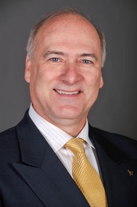 Headshot of Paul Kreider. He has gray hair and is smiling. He is wearing a navy blue jacket and a gold tie with a white, striped shirt. 