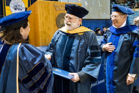 WVU 1983 School of Medicine  graduate Douglas B Learn, PhD walks and receives his diplomat 34 years later from WVU Provost Joyce McConnell, JD, LLM,  as friend Nyles Chardon PhD department of Microbiology looks on at WVU’s December commencement December 15, 2017.