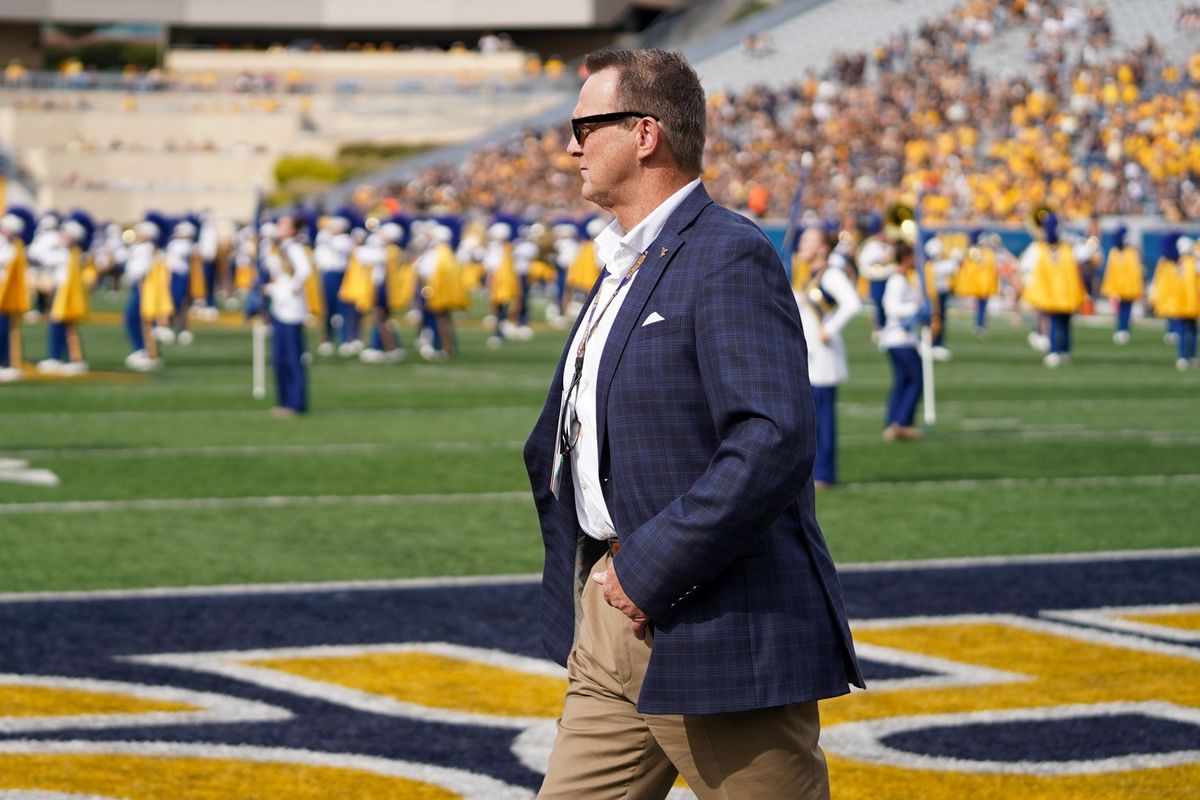 This is a photo of Shane Lyons walking the sideline at a WVU football game. He is wearing a navy blue suit jacket, white button up shirt and beige pants.