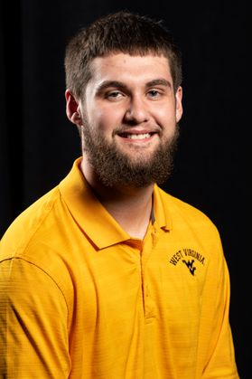 Photo of young man with dark hair and beard wearing gold shirt with flying WV below West Virginia against a dark background