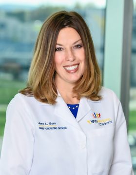 This is a portrait of Amy Bush who is standing in front of a large window. Bush has shoulder-length hair and is wearing a white doctor's coat with the logo for WVU Medicine Children's on the left side.