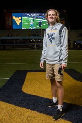 young man smiling, flying WV shirt with hood, standing with large screen in background
