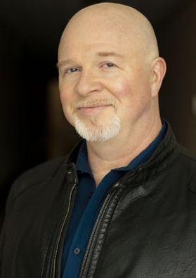 Headshot of WVU College of Creative Arts faculty member Lee Blair. He is pictured against a dark background wearing a black jacket over a dark blue shirt. He has a gray goatee 