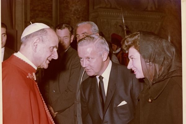 An old photo of a man in a red cape with a white hat talking to a man in a suit and woman in black wearing a scarf.