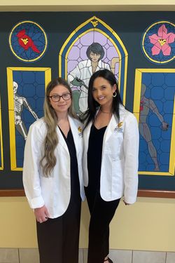 Photo of WVU dental students Savannah Lahey and Megan Merritt. They are pictured in their white lab coats and are standing in front of a mural wall. 