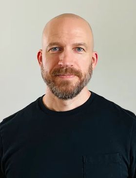 Headshot of WVU researcher William Franko. He is pictured standing in front of a light beige background. He is wearing a black shirt and has a shaved head and a full brown beard. 