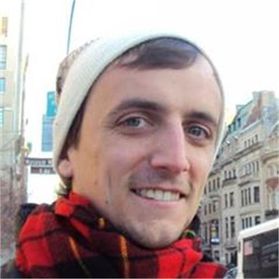Headshot of WVU professor Mason Moseley. He is pictured with a city scape behind him and is wearing a red plaid scarf and white winter hat. He has brown hair.