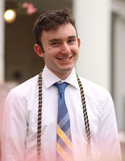 This is a portrait of Michael DiBacco who is wearing a white button-up shirt, light blue tie with a yellow plaid and a gold and blue cord. Michael has short, dark hair.
