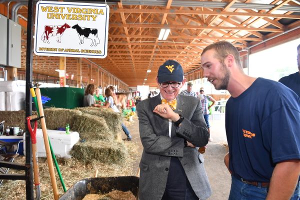 Two men standing in a barn. One man is wearing a blue hat with a gold WVU logo and a gray suite with a yellow bow tie. The other man is wearing a blue t-shirt and jeans.