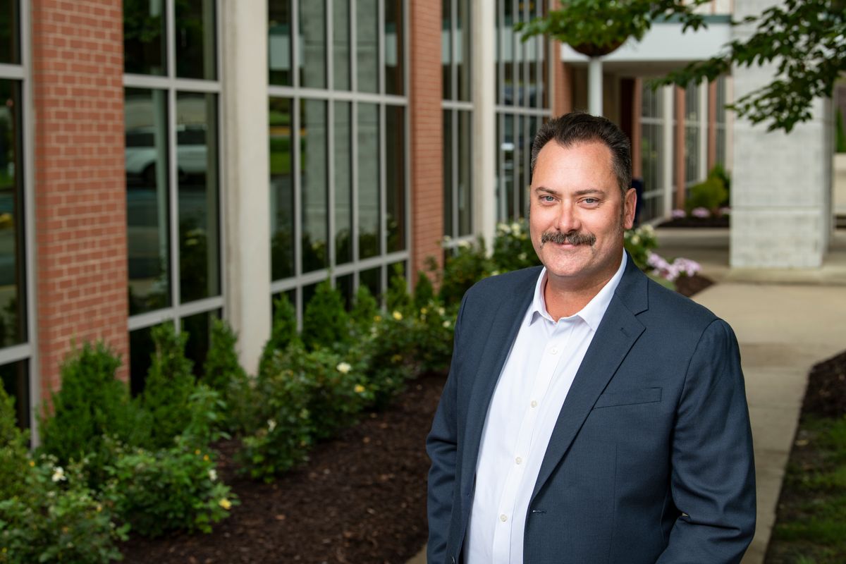 Community development expert Andy Williamson is pictured here standing in front of a building and wearing a dark suit with a white shirt. Williamson has dark hair and a mustache.