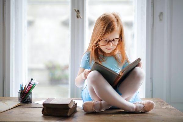 A small child with long golden blond hair sits on a desk while reading a book in front of a glass door.