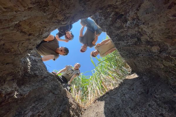 Photograph featuring graduate students looking down a deep hole for a research project. Five researchers are peering down into the hole with blue sky and greenery around them. 