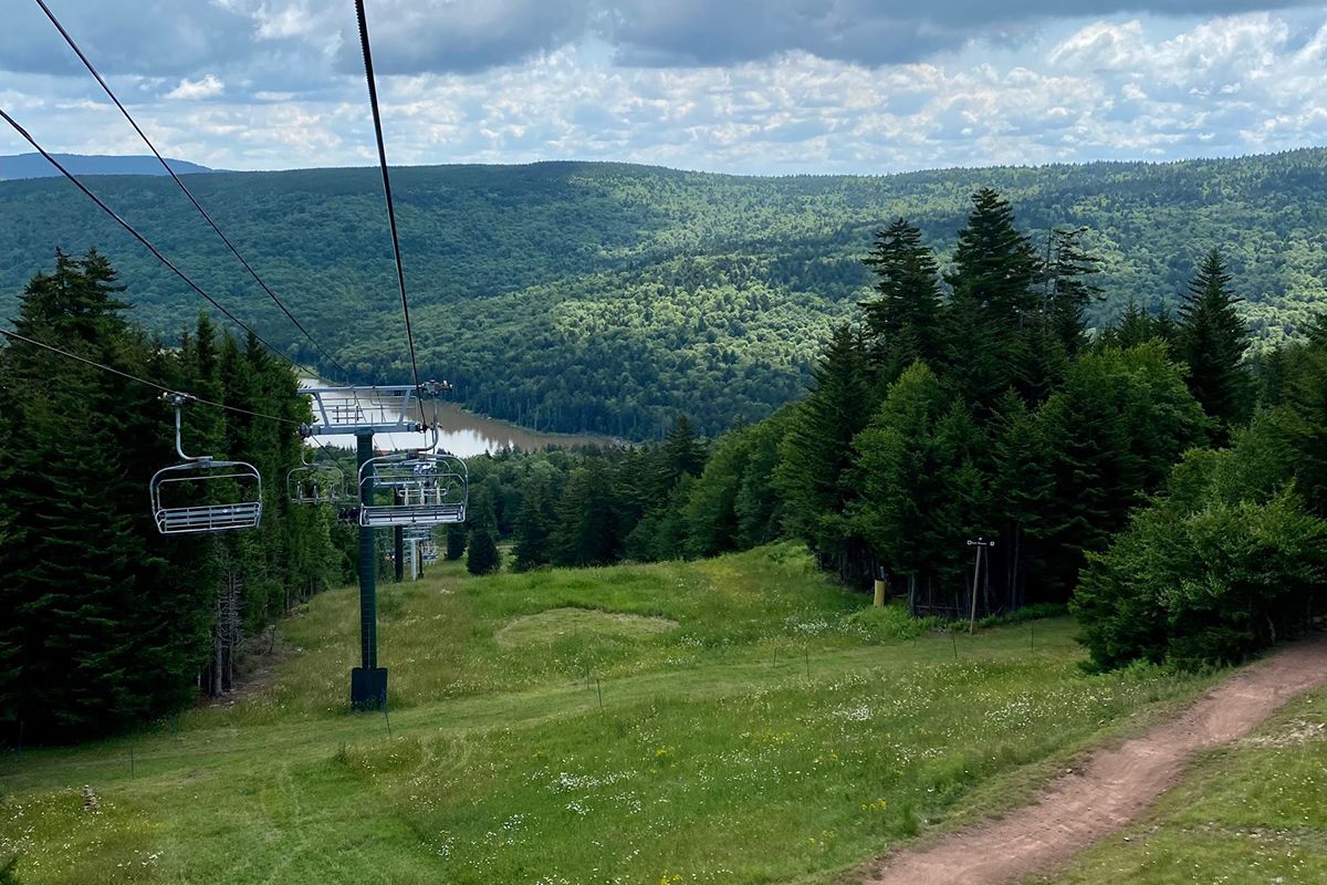 Lanscape photograph of Snowshoe Mountain Resort. A chair lift is pictured in the photo and you can see far into the valley below including the lake. The trees and grass are green. It is summer season at the resort. 