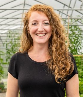 A person smiles while standing in front of plants in a greenhouse.