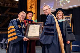 group of people in caps and gowns stands on a stage holding a framed diploma