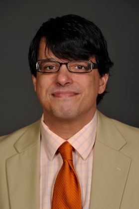 Man in tan suit jacket with orange and blue checkered button up shirt and an orange tie.