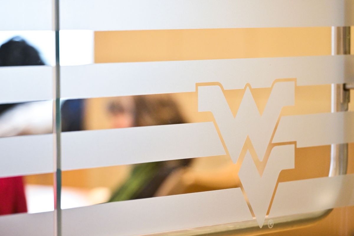 A window with a white Flying WVU sticker on it is shown here with a peek through the blinds into a room where people are working. 