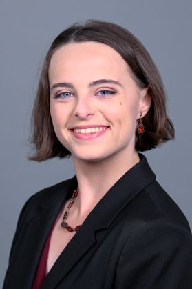 Headshot of WVU Bucklew Scholar Lillian Floyd. She is pictured against a gray background wearing a black jacket over a dark red shirt and a dark red necklace. She has brown hair cut into a bob just below her ears and is wearing red earrings. 