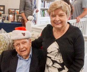 Picture of WVU donors Ken and Devon Gosnell during Christmas. Ken is seated on a couch wearing a dark blazer and a WVU Santa hat. His daughter, Devon, is seated next to him wearing a black and cream floral dress. They are both elderly. 