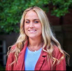 Headshot of WVU student Hillary Probst. She is standing outside with greenery behind her. She is wearing a red jacket over a light blue shirt. She has long blonde hair.