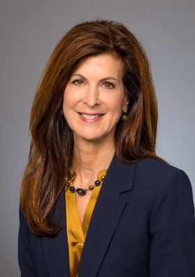 Headshot of WVU administrator Paula Congelio. She is pictured in front of a gray background wearing a navy blue jacket over a gold satin blouse. She has long, auburn colored hair. 