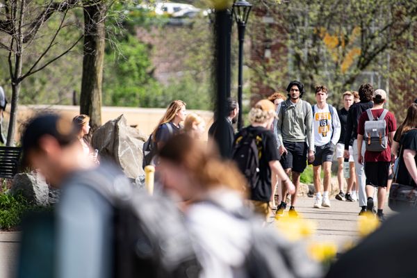 More than a dozen people, many wearing backpacks, walk down sidewalks on WVU's Morgantown campus. Trees are visible in the background.