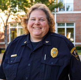 WVU Police Chief Sherry St. Clair is pictured standing outside with a red brick building behind her. She is wearing a navy blue uniform and has a gold badge on her left breast pocket. She has shoulder length wavy reddish hair. 
