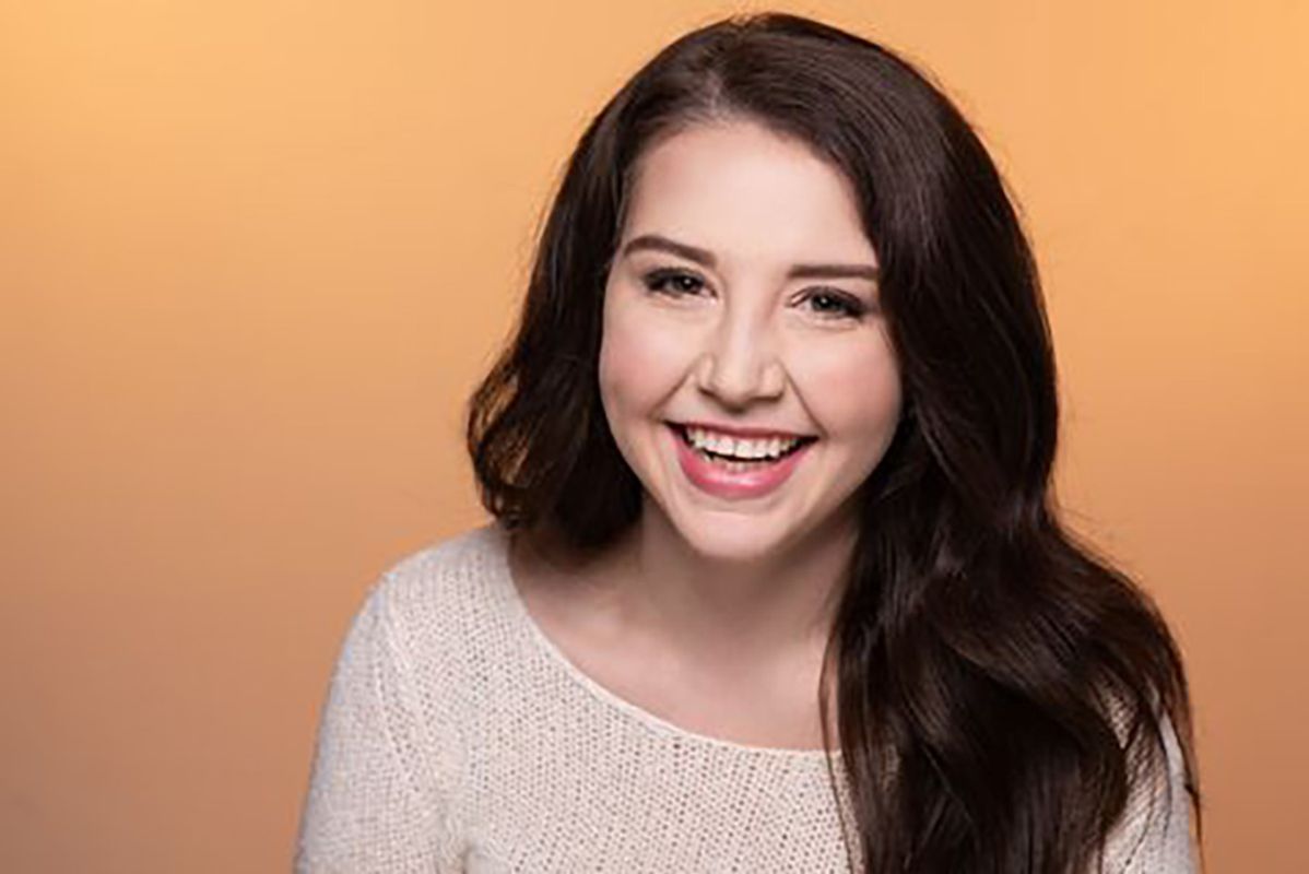 Headshot of WVU MFA student Josey Miller. She is pictured against a peach colored background wearing a cream colored sweater. She has long, brown hair.
