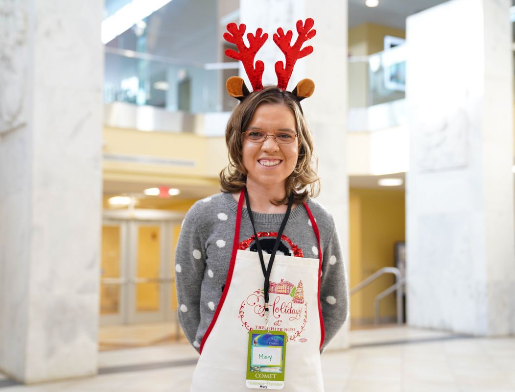 Photograph of WVU employee Mary Veselicky. She is standing in the lobby of the WVU Health Science buildings with three large white marble columns in the background. She is wearing red reindeer antlers and a festive apron. 