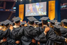 Graduates are shown from the back with their arms linked during Commencement at the Coliseum. Gold Commencement banners are shown in the background of the photo.